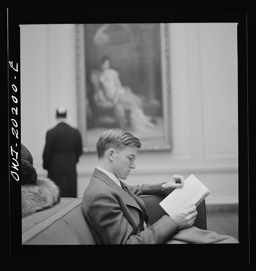 Black and white picture of a man sitting in an art gallery and reading a book